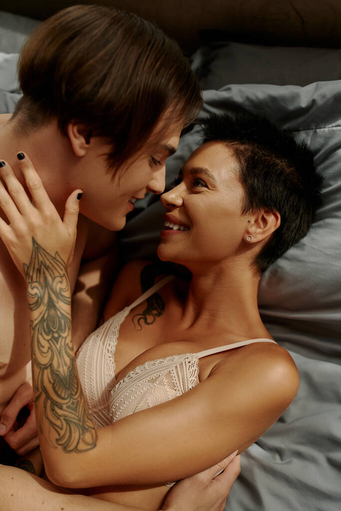 Χαμογελαστή γυναίκα με τατουάζ στο σουτιέν να αγγίζει τον γυμνό φίλο της στο κρεβάτι.  - Φωτογραφία, εικόνα