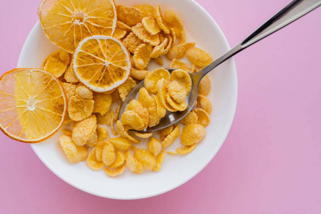 верхний вид сушеных апельсинов в миске с кукурузными хлопьями и ложкой на розовом фоне  - Фото, изображение