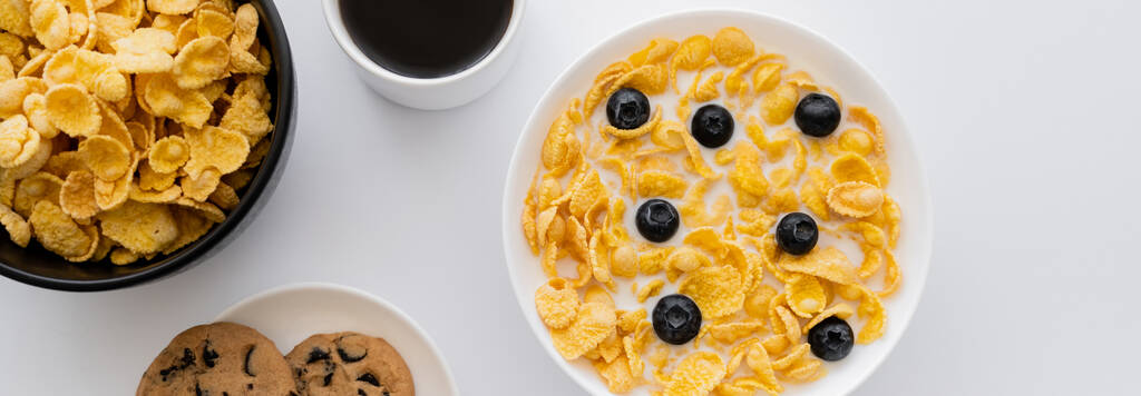 shora pohled na misky s kukuřičnými vločkami v mléce s borůvkami u šálku kávy a sušenky izolované na bílém, banner - Fotografie, Obrázek