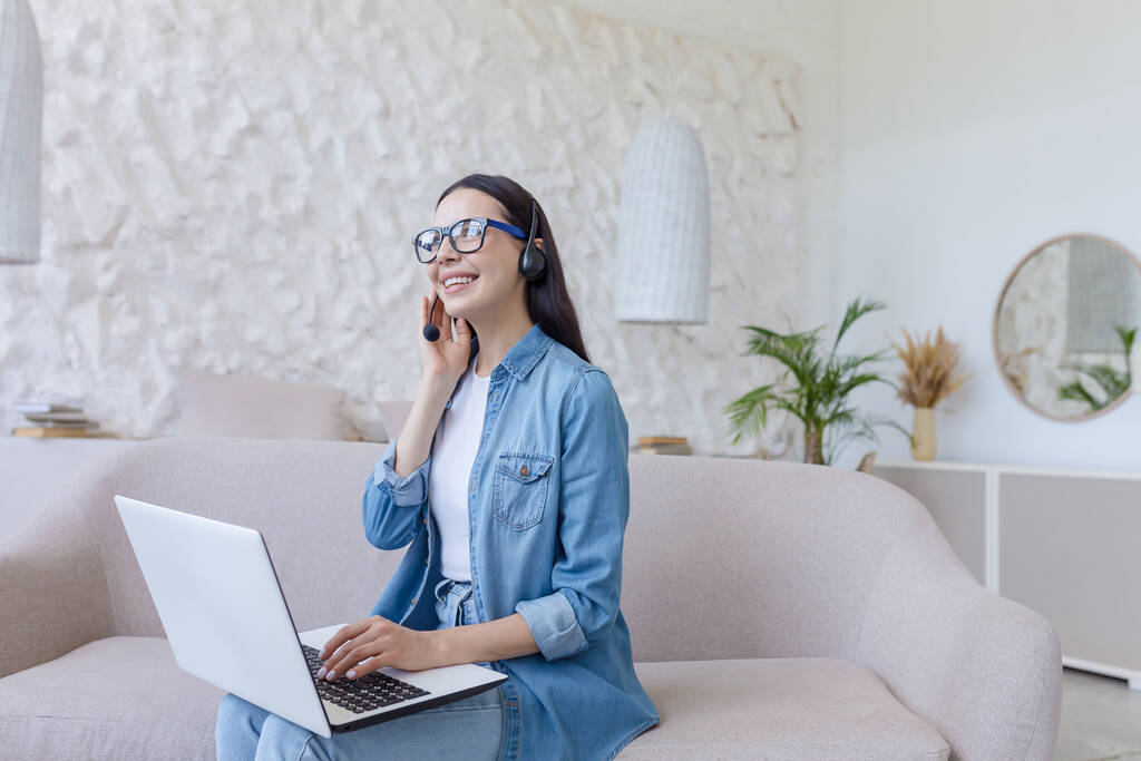 Online work. Young beautiful freelancer woman in glasses and jeans sitting at home on sofa and working with laptop and headphones online, talking on video call with clients, partners. - Photo, image