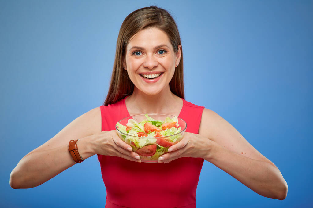 Jonge lachende dame in rode jurk met salade kom geïsoleerd portret op blauwe achtergrond. - Foto, afbeelding