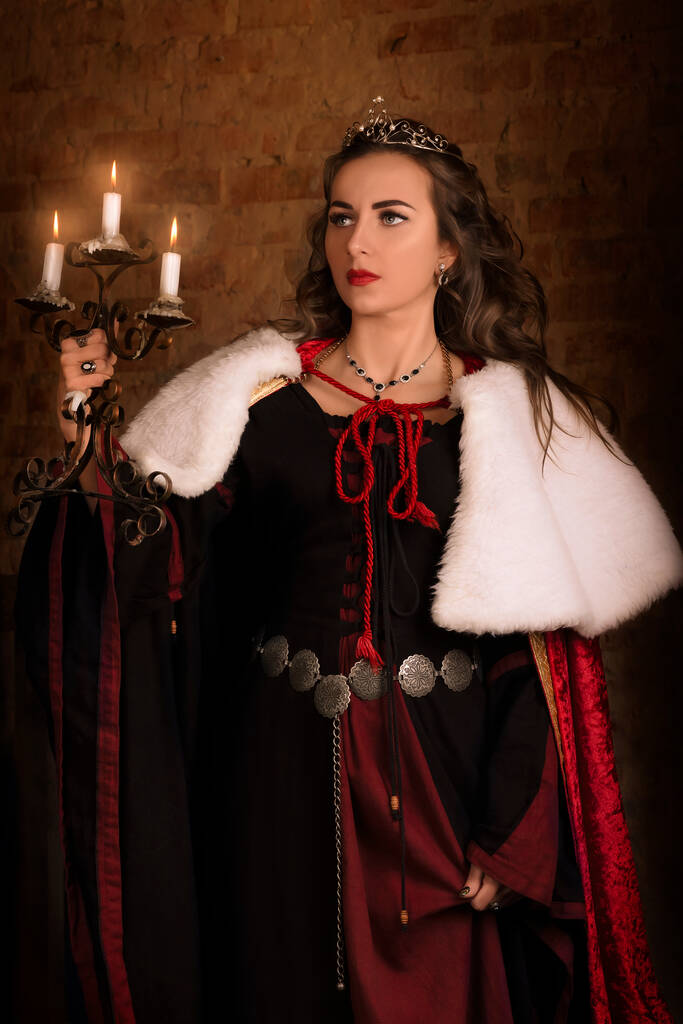 Fantastische middeleeuwse vrouw met een kandelaar in haar handen. Royal fluwelen vintage rode jurk met bont en goud, prinses, koningin, brunette met lang haar. Kasteel, schemering, nacht, luxe - Foto, afbeelding