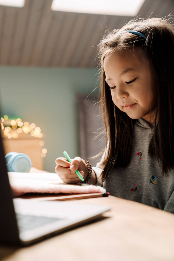 Aziatisch meisje doet huiswerk met laptop terwijl zitten aan tafel thuis - Foto, afbeelding