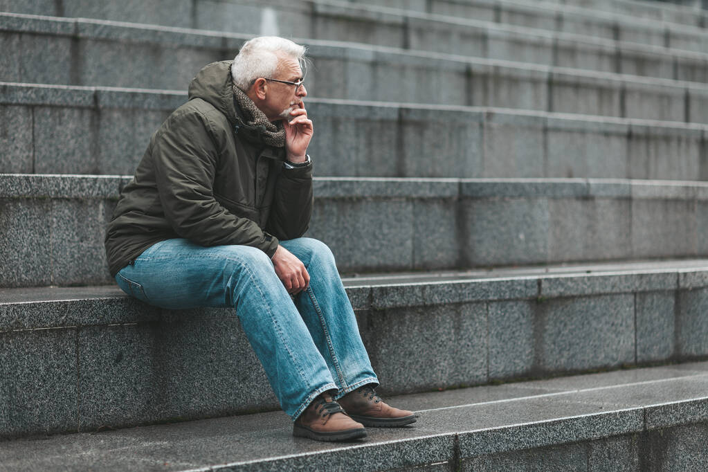 El hombre de pelo gris está perdido en el pensamiento y se sienta en las escaleras. Nostalgia de un pensionista. - Foto, Imagen