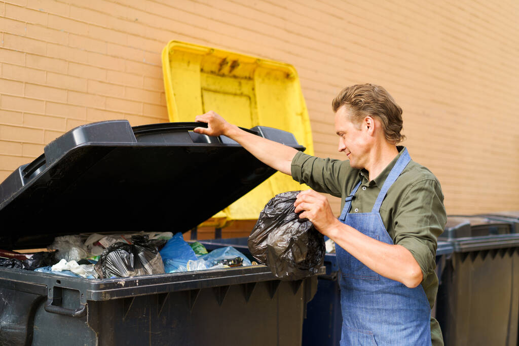 Caucasian handsome man employee open can full of garbage to throw plastic bag with trash wearing apron. Wrong unsorted garbage cans. Wrong way to throw your trash. Sorting garbage save the world.  - Photo, Image