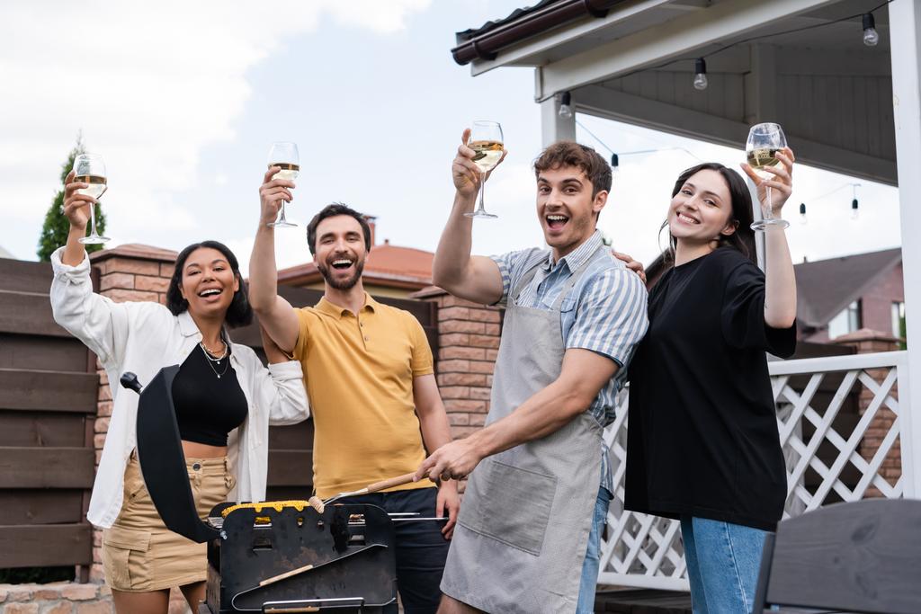 Excited multiethnic friends holding wine and grilling corn and meat outdoors  - Photo, Image