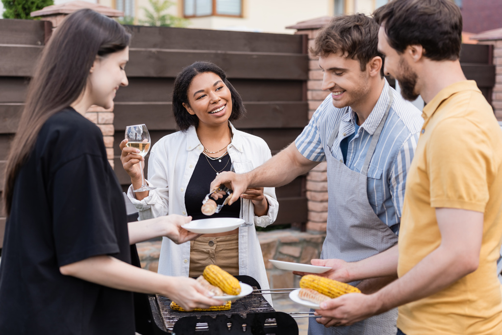 Hombre en delantal poniendo comida a la parrilla en platos cerca de amigos multiétnicos con vino  - Foto, Imagen