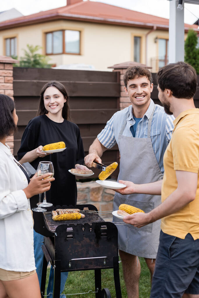 Hymyilevä mies laittaa grillattua maissia lautaselle lähellä monikansallisia ystäviä viinin kanssa takapihalla  - Valokuva, kuva