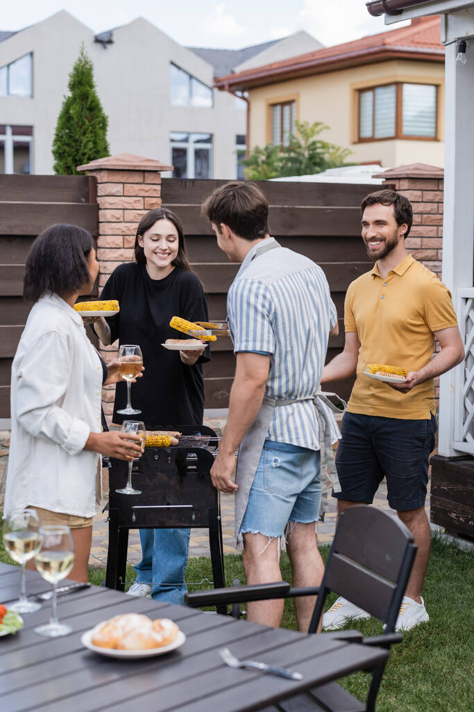 Amici multietnici sorridenti con vino e cibo su piatti in piedi vicino alla griglia nel cortile  - Foto, immagini