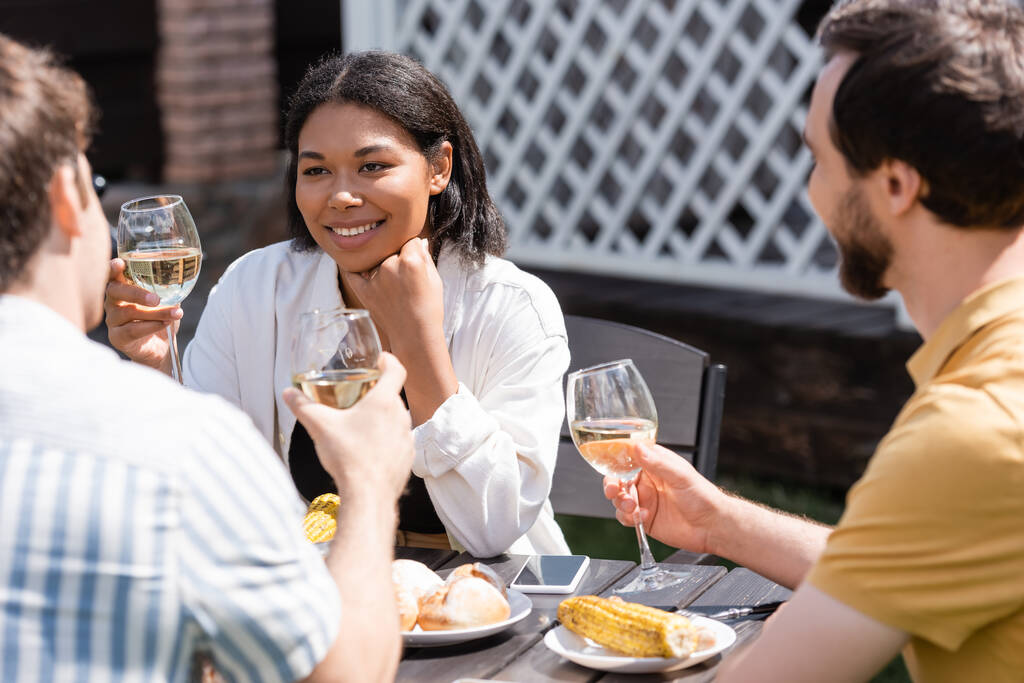 Улыбающаяся двурасовая женщина с бокалом вина рядом с размытыми друзьями и едой на заднем дворе  - Фото, изображение