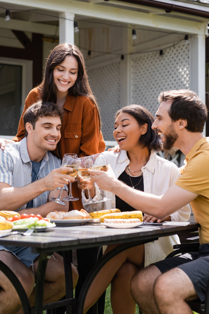 Allegri amici multietnici brindano con vino vicino al cibo alla griglia durante il picnic all'aperto  - Foto, immagini