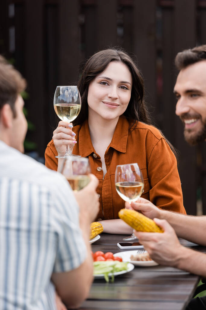 Mujer joven sosteniendo una copa de vino y mirando a la cámara cerca de amigos borrosos durante el picnic al aire libre  - Foto, imagen