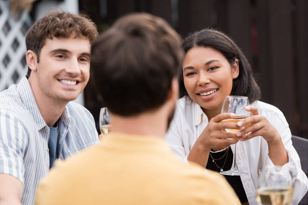 feliz pareja multiétnica con copas de vino mirando a un amigo en primer plano borroso - Foto, imagen