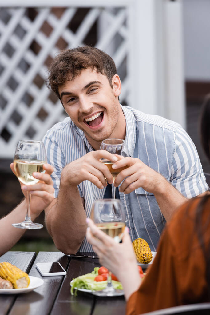 hombre emocionado sosteniendo copa con vino cerca de amigos durante la fiesta de barbacoa en el patio trasero  - Foto, imagen