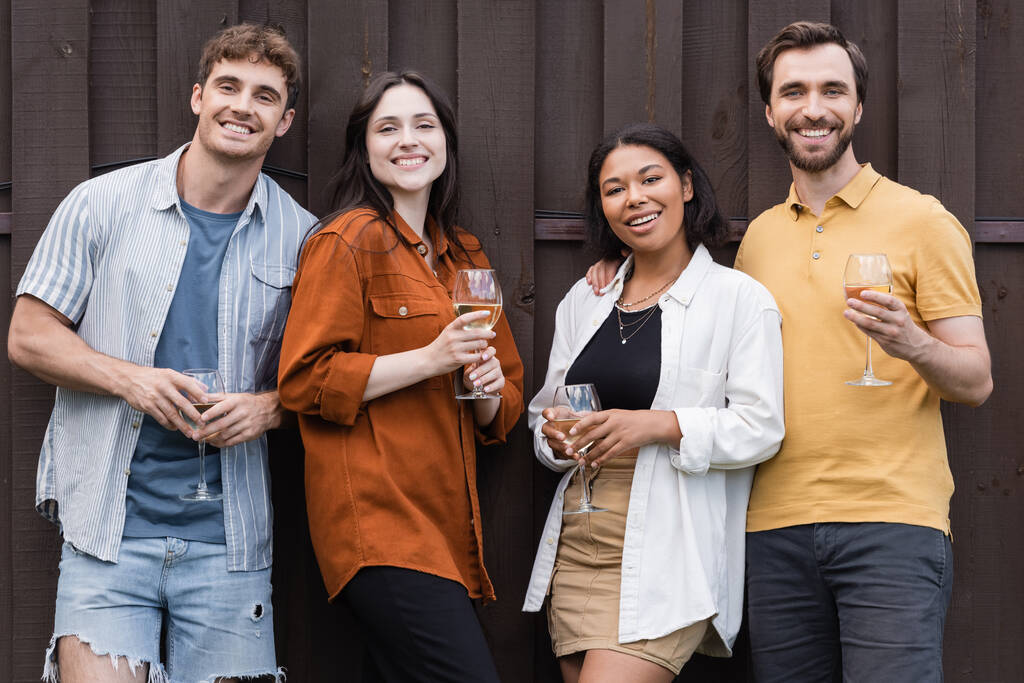 happy interracial friends holding glasses with wine while standing near fence on backyard - Photo, Image