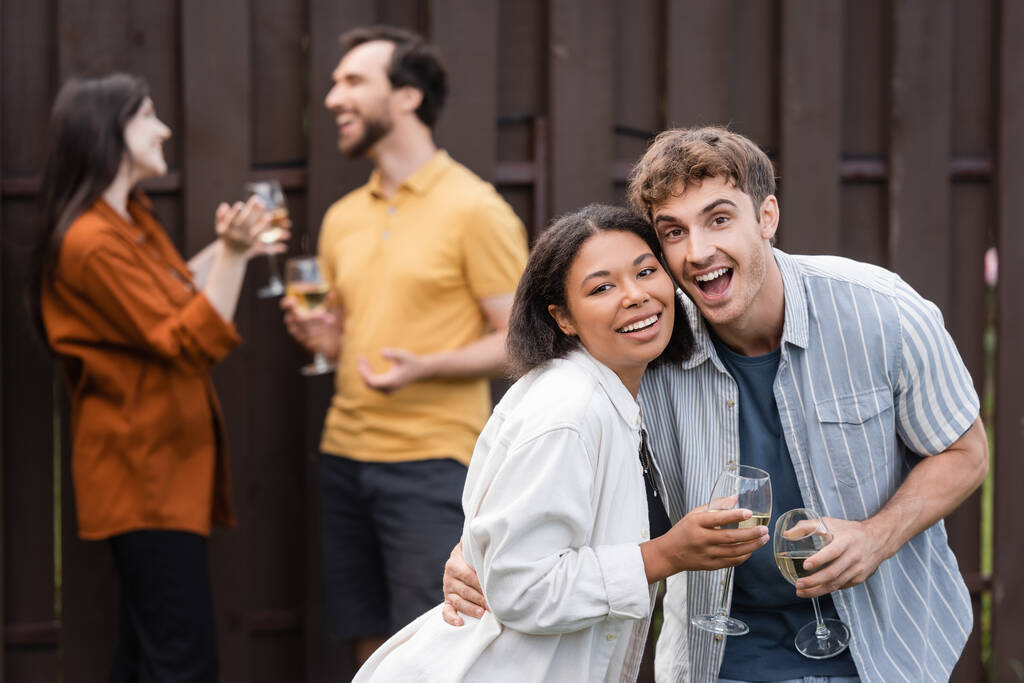 alegre interracial pareja celebración vasos con vino cerca borrosa amigos en borrosa fondo - Foto, Imagen