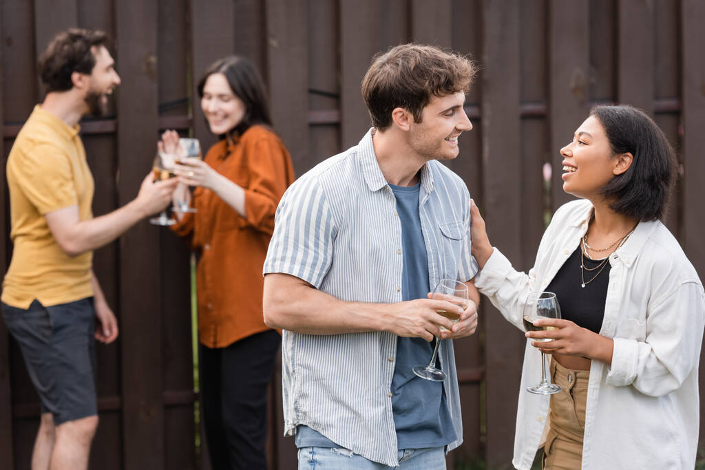 veselý mezirasový pár drží sklenice s vínem, zatímco mluví v blízkosti rozmazaných přátel na rozmazaném pozadí - Fotografie, Obrázek