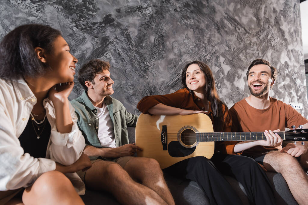 fröhliche Frau mit Akustikgitarre neben multiethnischen Freunden, die auf der Couch im modernen Wohnzimmer sitzen  - Foto, Bild