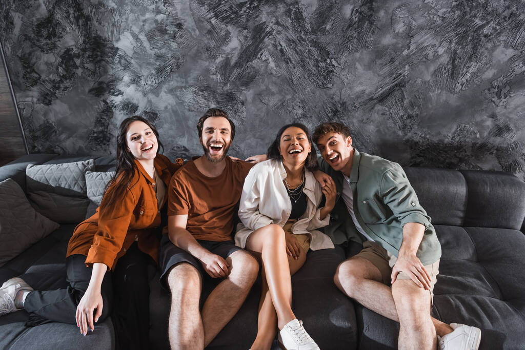 χαρούμενους πολυπολιτισμικούς φίλους με casual ρούχα που κάθονται σε γκρι καναπέ στο μοντέρνο σαλόνι - Φωτογραφία, εικόνα