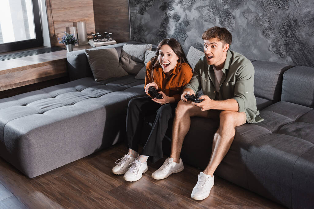 KYIV, UCRANIA - 26 de julio de 2022: amigos emocionados sosteniendo joysticks y jugando videojuegos mientras están sentados en un sofá gris - Foto, Imagen
