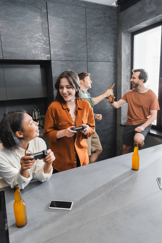 KYIV, UCRANIA - 26 de julio de 2022: alegres mujeres interracial jugando videojuegos cerca de amigos en la cocina  - Foto, imagen