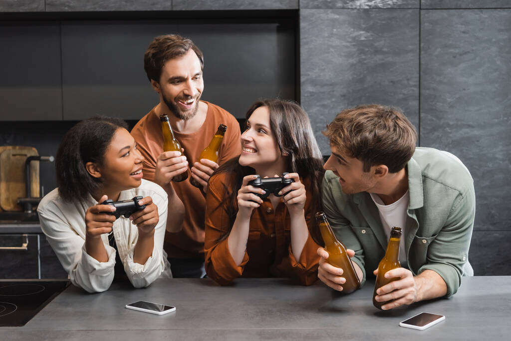 KYIV, UCRANIA - 26 de julio de 2022: mujeres interracial felices sosteniendo joysticks cerca de amigos varones con cerveza en la cocina  - Foto, imagen