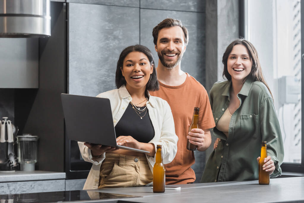 щаслива бі-расова жінка тримає ноутбук поруч з друзями з пляшками пива, що стоять на кухні
  - Фото, зображення