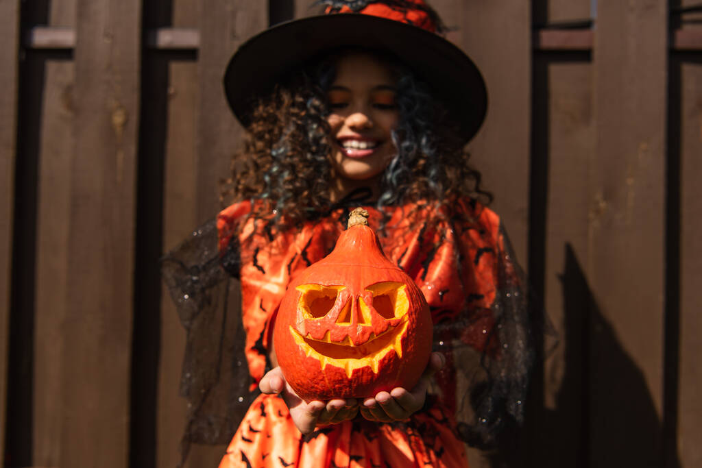 foco seletivo de abóbora esculpida em mãos de menina sorridente em traje de bruxa halloween - Foto, Imagem