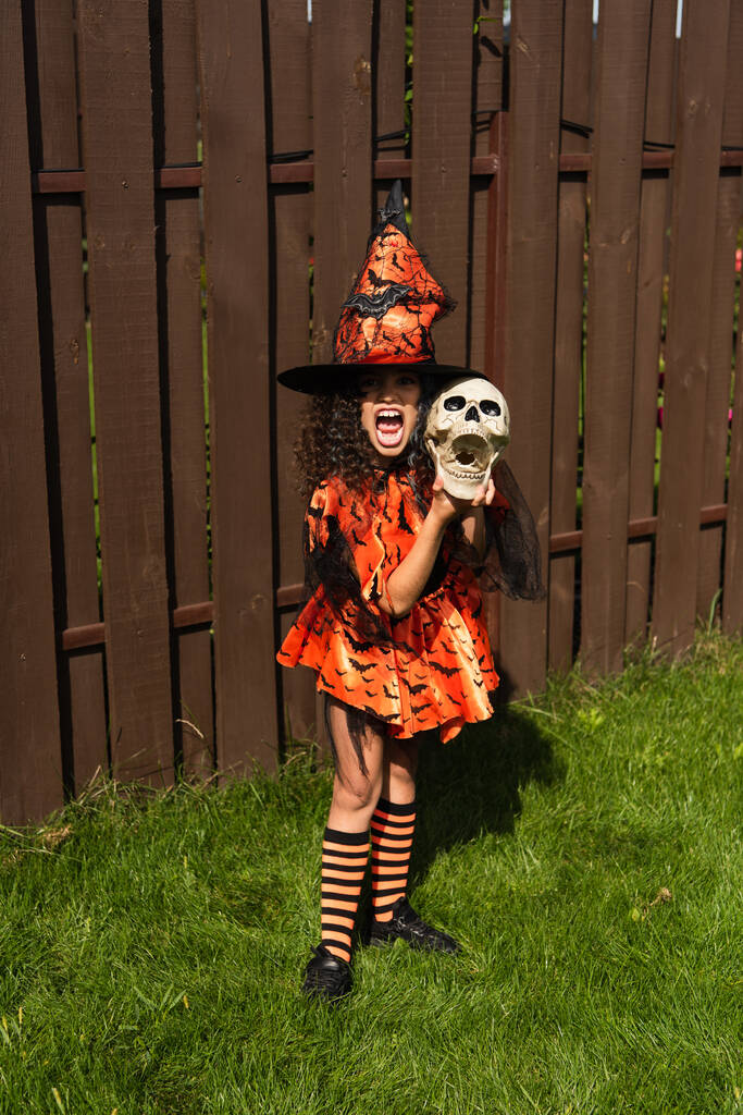 Mädchen in voller Länge in Halloween-Kostüm und Hexenhut mit gruseligem Totenkopf und Grimassen im Freien - Foto, Bild