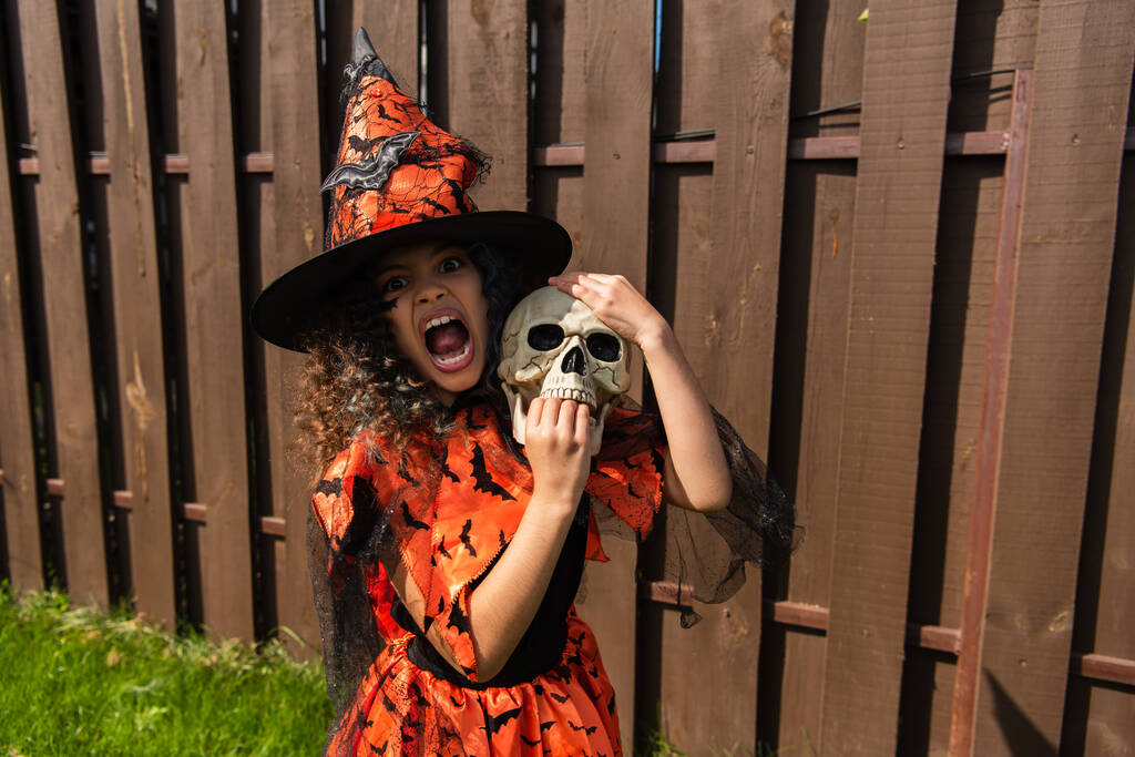 enfant en costume de sorcière tenant crâne effrayant, grimaçant et grognant à l'extérieur - Photo, image