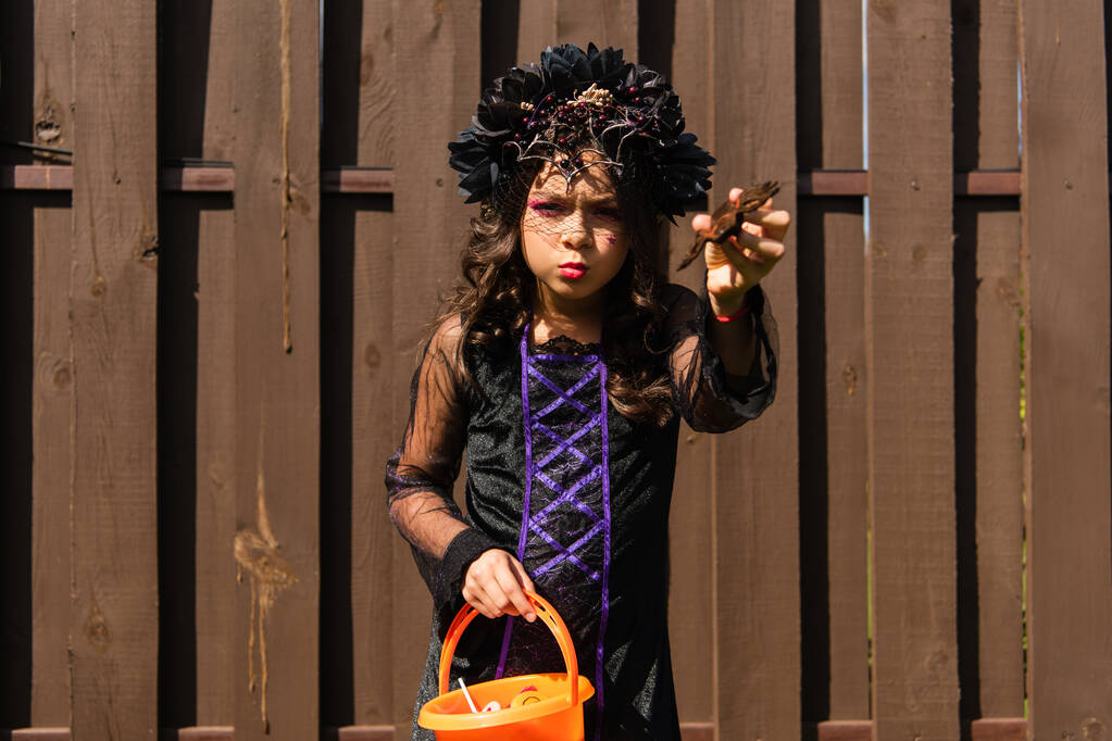 Mädchen im Hexenkostüm hält Halloween-Eimer und erschreckt mit Spielzeugspinne - Foto, Bild