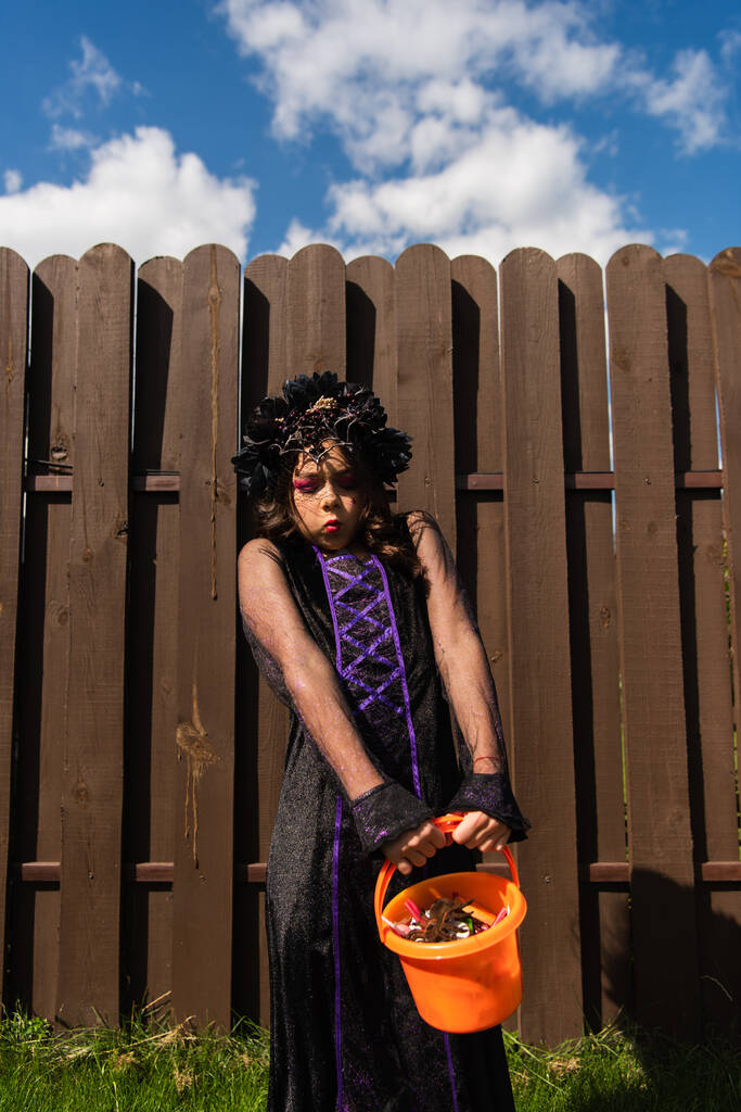 dívka v kostýmu čarodějnice a černý věnec drží těžký kbelík s bonbóny - Fotografie, Obrázek