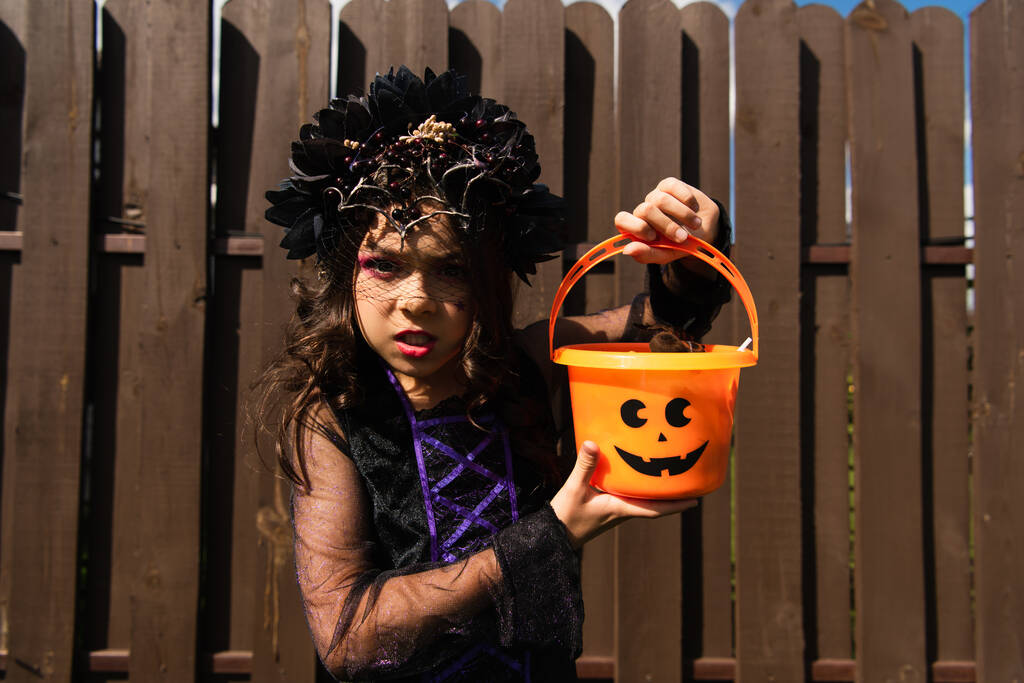 Cadılar Bayramı kovası tutan ve kızgın suratıyla kameraya bakan cadı kostümlü bir kız. - Fotoğraf, Görsel