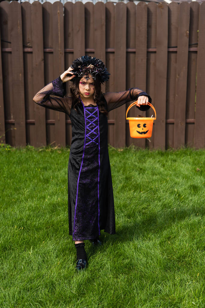 serious girl in black dress and wreath holding halloween bucket while walking in backyard - Photo, Image