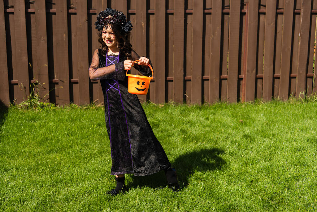 longitud completa de la muchacha en traje de bruja caminando con cubo de Halloween en el césped en el patio trasero - Foto, imagen