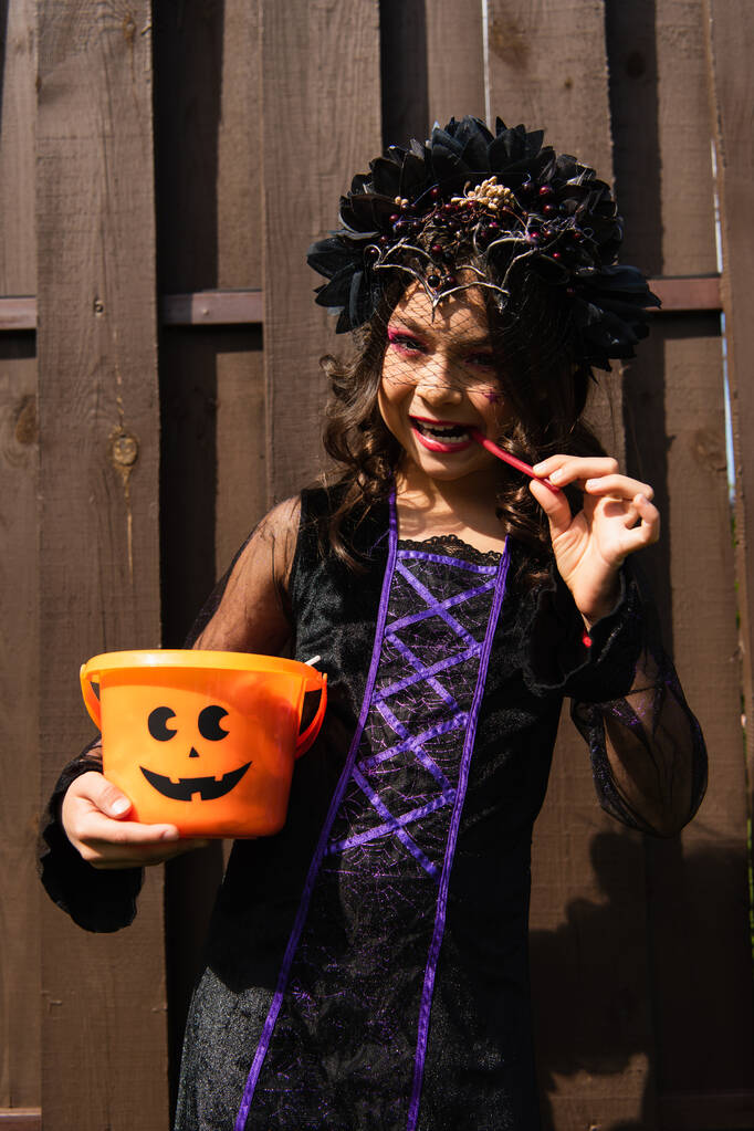 dívka v kostýmu čarodějnice drží halloween vědro a jíst lékořice hůl - Fotografie, Obrázek