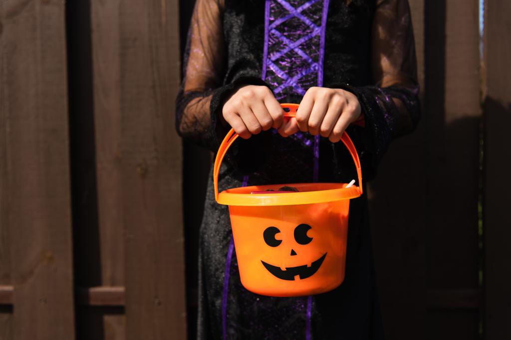 abgeschnittene Ansicht eines Mädchens im Hexenkostüm, das einen Halloween-Eimer im Freien hält - Foto, Bild