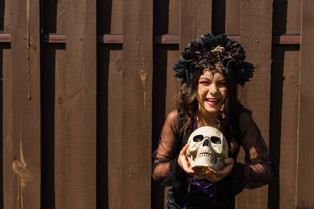 wesoła dziewczyna w czarnym wieńcu trzyma straszną czaszkę i uśmiecha się do kamery w pobliżu drewnianego ogrodzenia - Zdjęcie, obraz