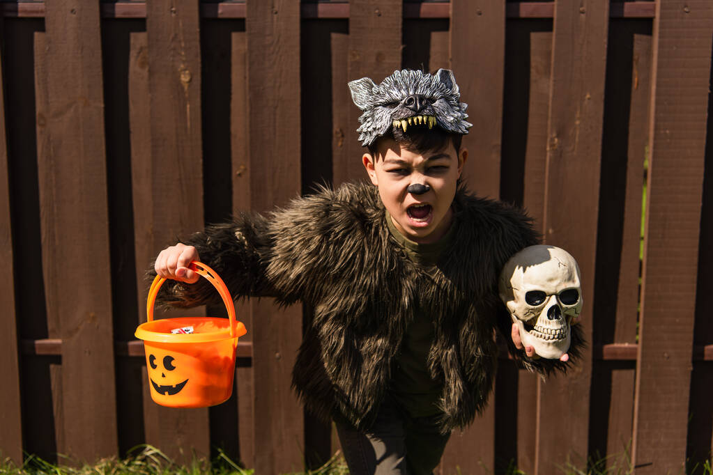 asiático chico en hombre lobo traje corriendo con cráneo y truco o tratar cubo mientras miedo gritando al aire libre - Foto, Imagen