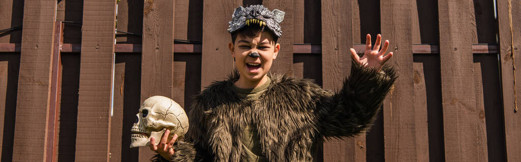asiatischer Junge im Werwolf-Kostüm mit wütender Fratze und beängstigender Geste, während er einen gruseligen Totenkopf hält, Banner - Foto, Bild