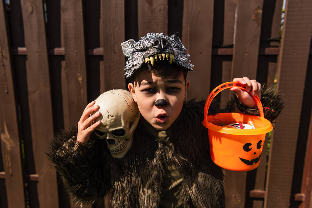 aufgeregter asiatischer Junge im Werwolf-Kostüm mit Totenkopf und Halloween-Eimer mit Süßigkeiten - Foto, Bild