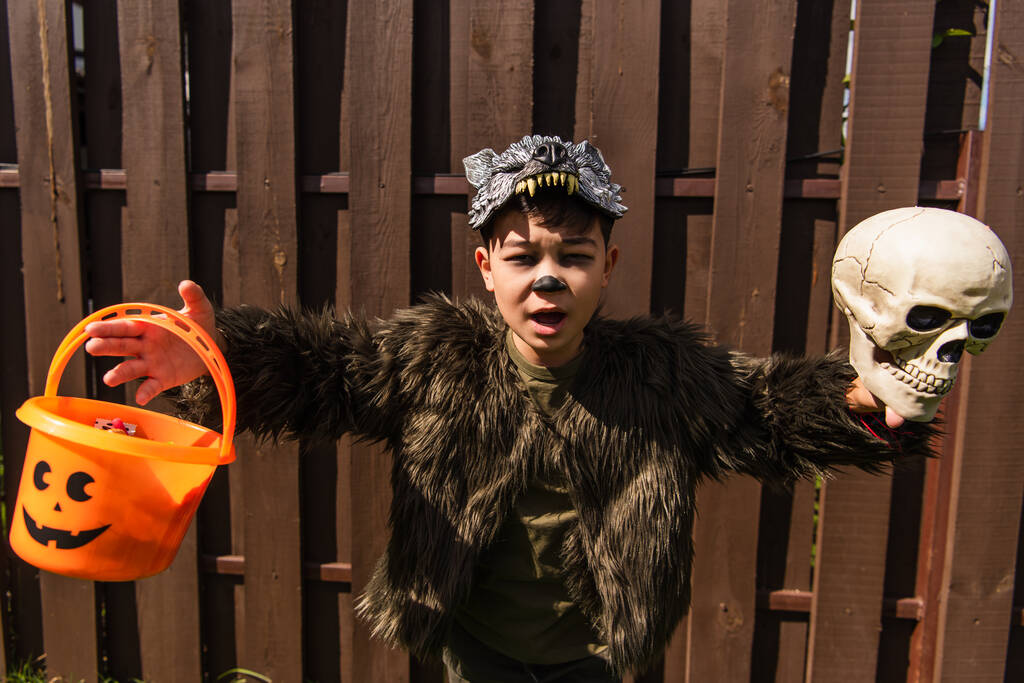 espantado asiático menino no lobisomem traje correndo com crânio e halloween balde no quintal - Foto, Imagem