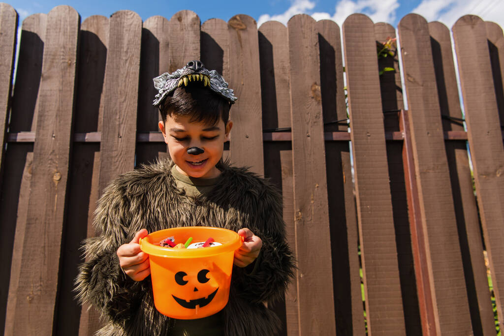 zadowolony azjatycki chłopiec w wilkołak kostium patrząc w Halloween wiadro ze słodyczami - Zdjęcie, obraz