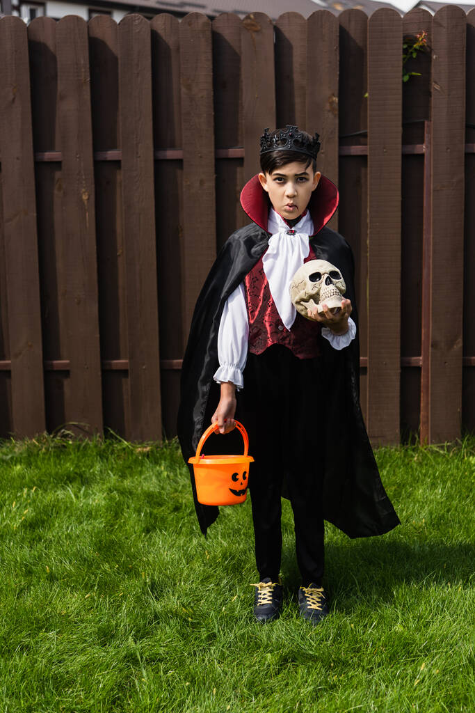 ázsiai fiú vámpír király jelmez és dühös grimasz holding halloween vödör és koponya - Fotó, kép