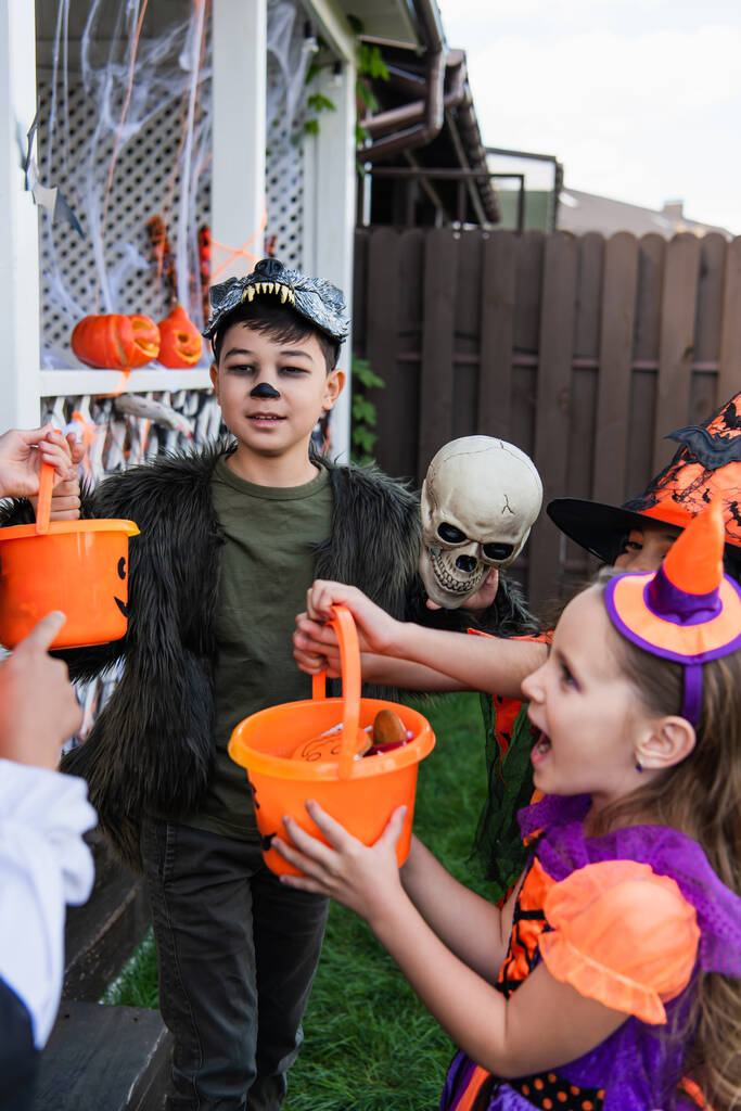 Asiatico ragazzo in halloween costume holding secchio con caramelle vicino amico in cortile  - Foto, immagini