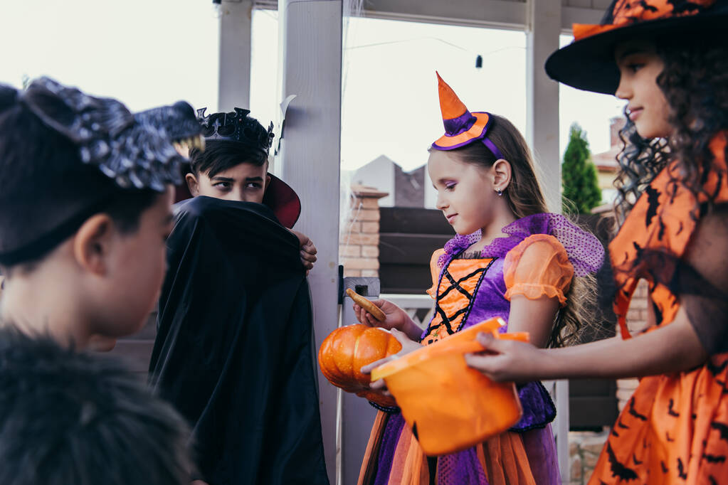 Ребенок держит тыкву и печенье рядом с размытыми многонациональными друзьями в костюмах на Хэллоуин на открытом воздухе  - Фото, изображение