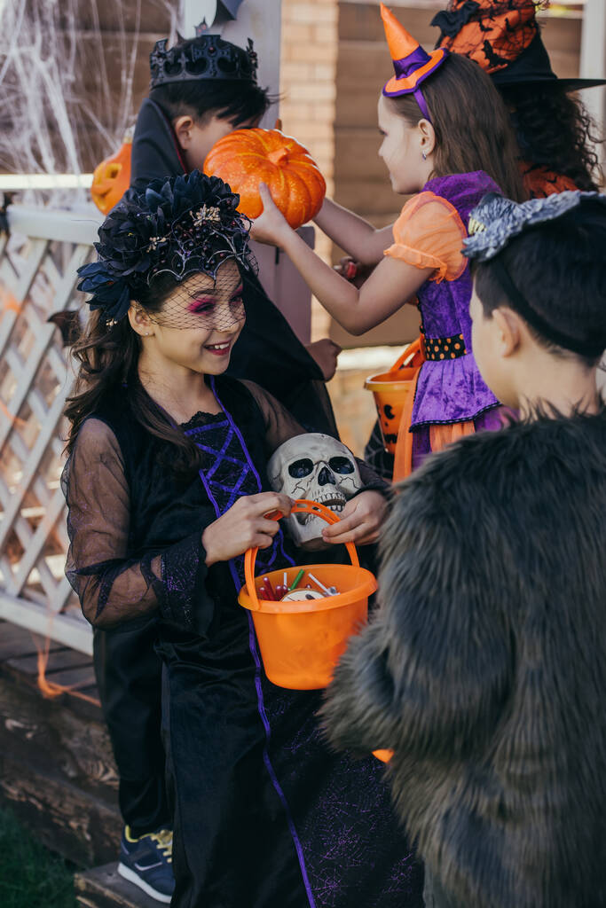 Enfant gai en costume d'Halloween tenant le crâne et le seau avec des bonbons près d'un ami asiatique à l'extérieur  - Photo, image