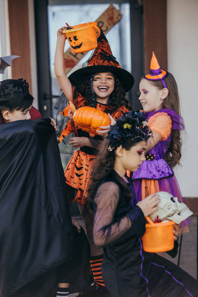 Wesoła dziewczyna w kapeluszu czarownicy trzymając wiadro w pobliżu wielonarodowych przyjaciół podczas obchodów Halloween na świeżym powietrzu  - Zdjęcie, obraz