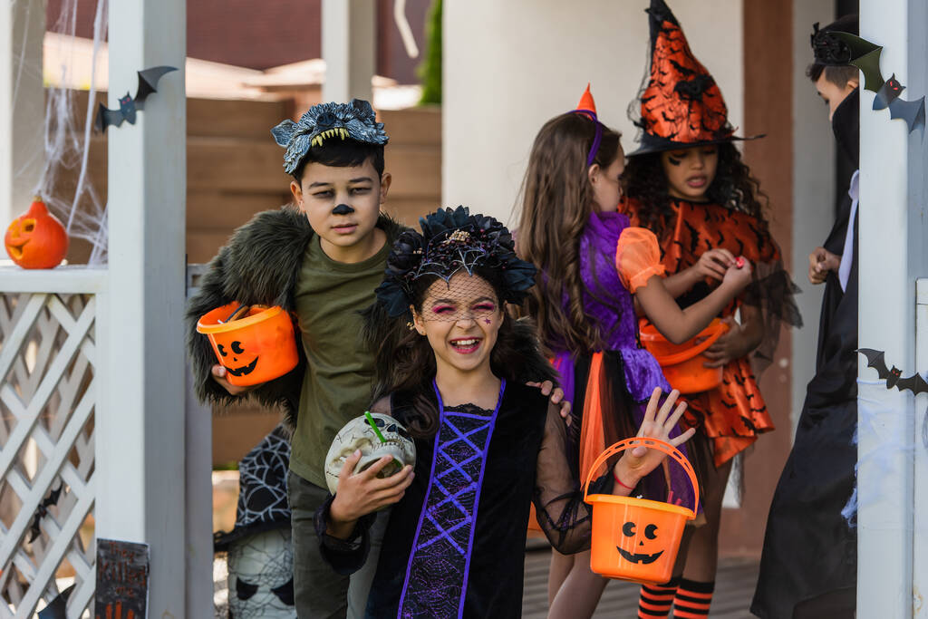 Ragazza felice in costume di Halloween tenendo teschio vicino amico asiatico con secchio e bambini in cortile  - Foto, immagini