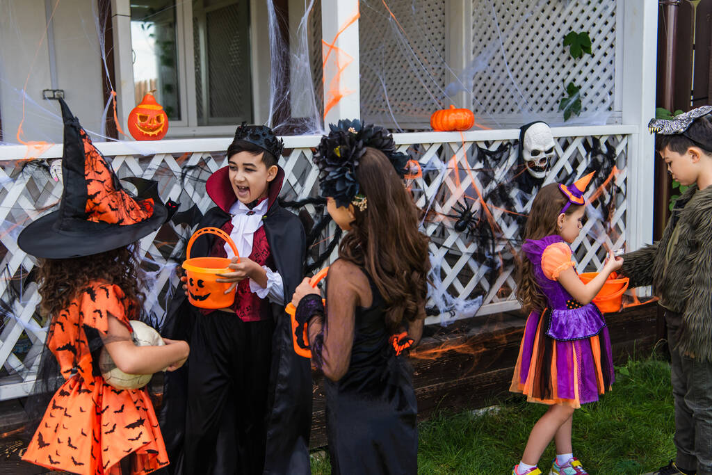 Enfants multiethniques en costumes d'Halloween tenant des seaux près de toile d'araignée et décor sur la maison  - Photo, image